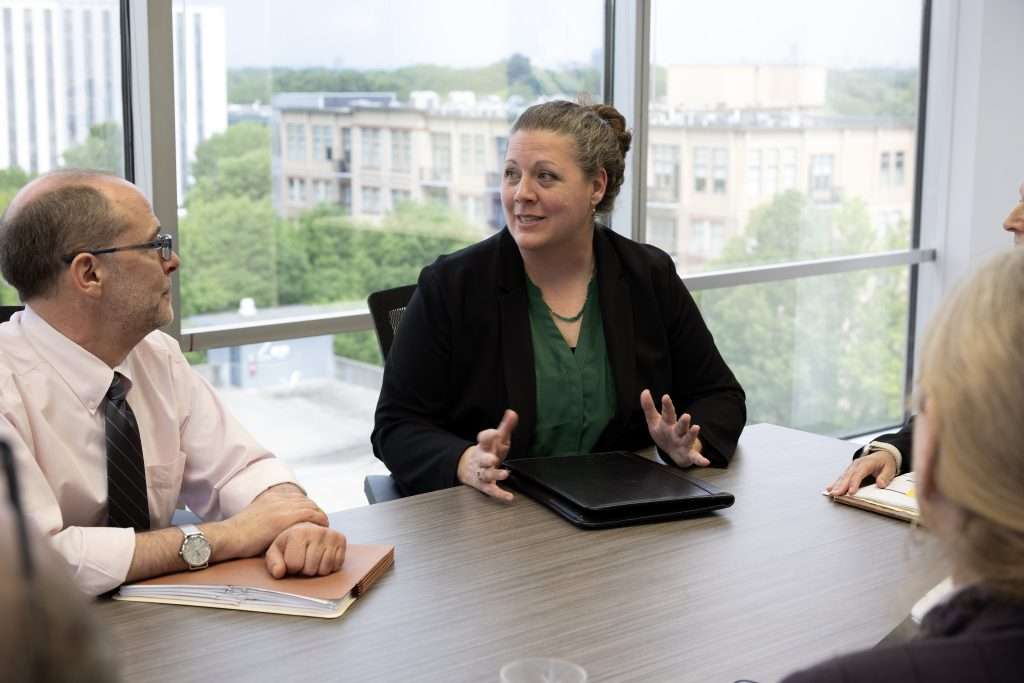 Kristen Howe wearing a black blazer and green shirt, sits at a conference table with three colleagues, holding a discussion. The background shows large windows with a view of buildings and trees.