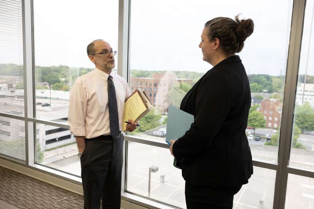 Kristen Howe stands in an office beside a large window with a cityscape view. She faces the other person, who holds a document and is also dressed in business clothing. They appear to be engaged in conversation.