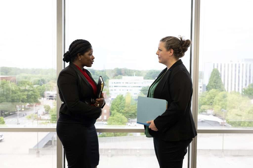 Two professional women stand facing each other in front of a large window with a cityscape and greenery visible outside. They are engaged in conversation, each holding folders and wearing business attire.