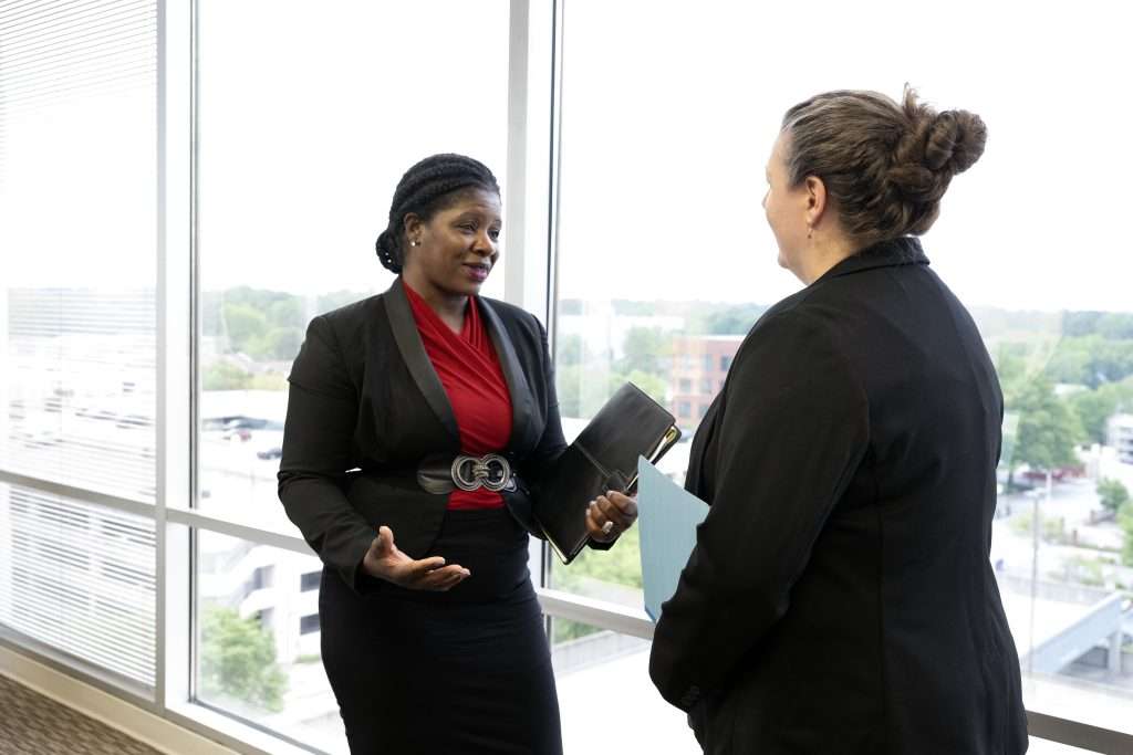Two women in business attire are engaged in a conversation by a large window with a cityscape and greenery in the background. One woman is holding a black portfolio, while the other has a folder. They appear to be in an office setting.